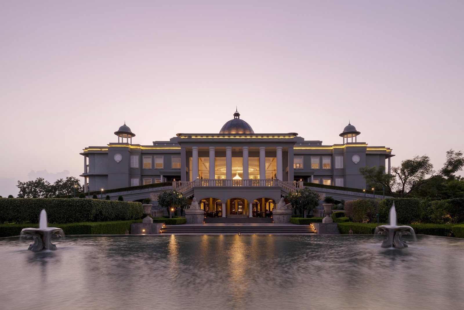 Raffles Udaipur at dusk, its lit colonnaded facade mirrored in the water below
